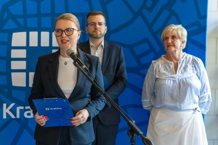 Kobieta w garniturze przemawia do mikrofonu, w tle mężczyzna i starsza kobieta.