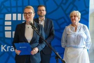 Kobieta w garniturze przemawia do mikrofonu, w tle mężczyzna i starsza kobieta.