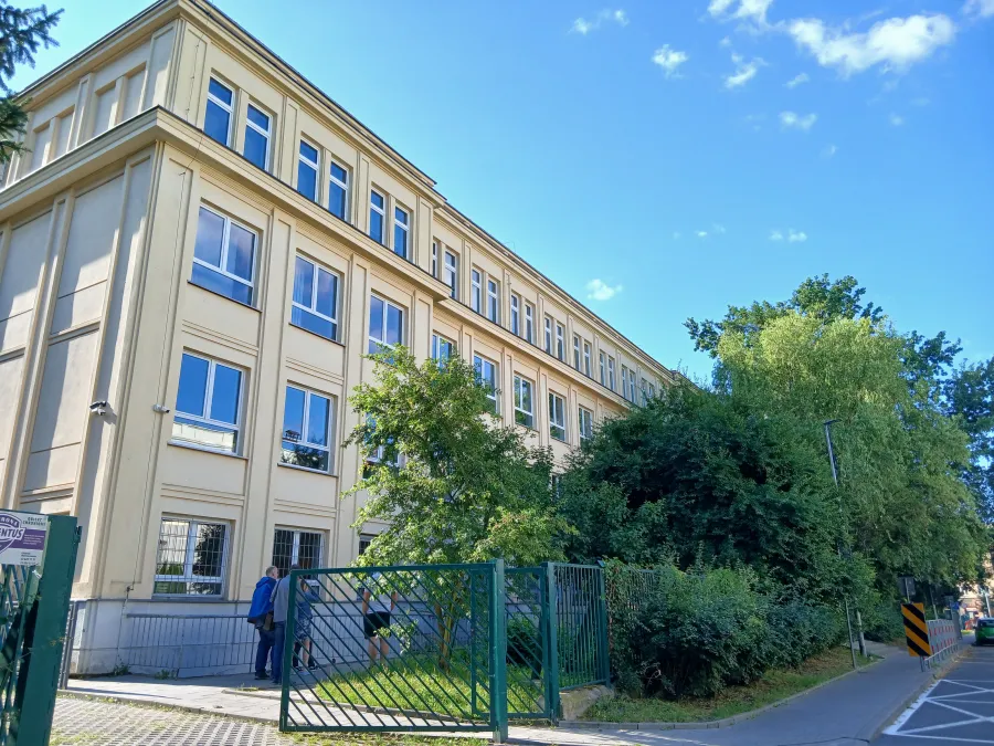 Budynek z dużymi oknami otoczony zielenią, widok z ulicy.