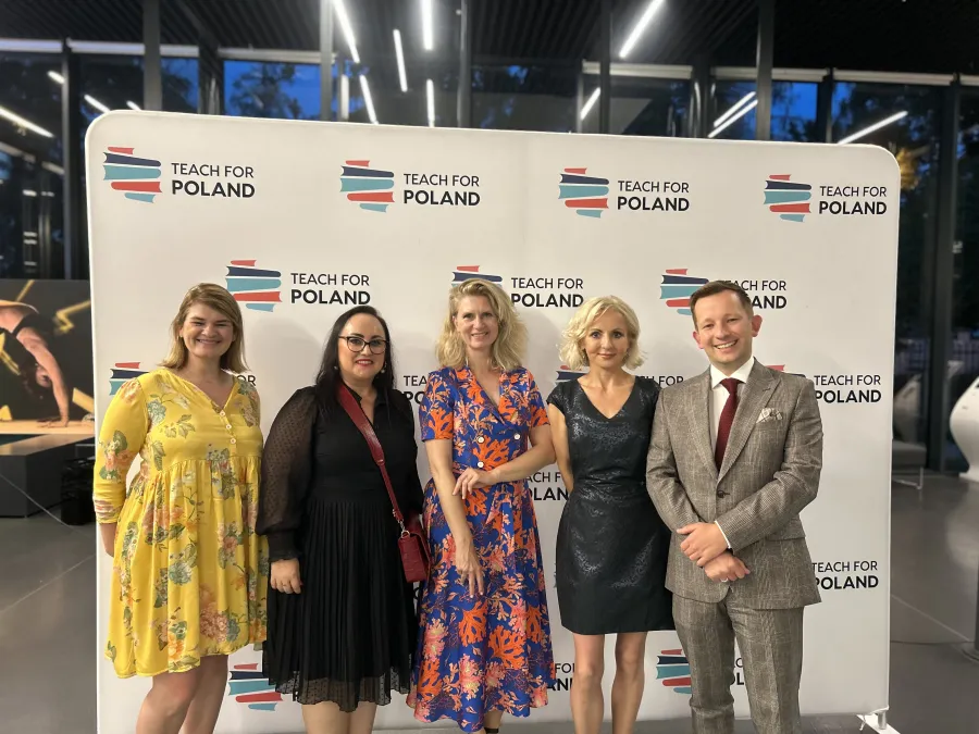 Grupa pięciu osób w eleganckich strojach na tle banera Teach for Poland.