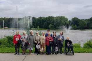 Grupa osób starszych pozujących nad jeziorem w parku. W tle fontanny i zieleń.