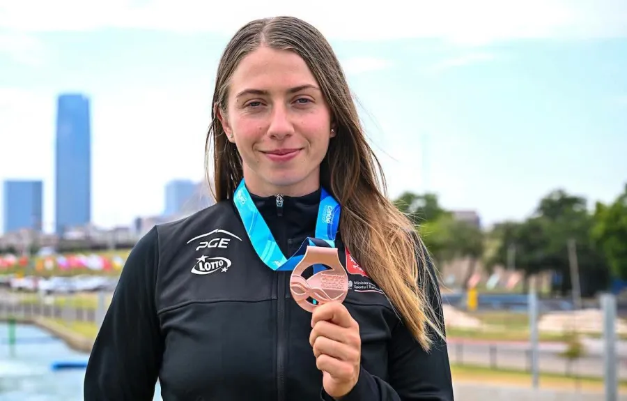 Sportowiec trzyma brązowy medal, uśmiechając się w tle z widokiem na miasto.