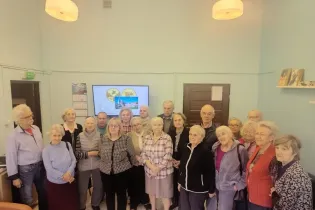 Grupa seniorów w sali, z ekranem w tle i kalendarzem na ścianie.