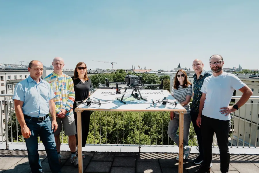 Grupa ludzi stojąca przy stole z dronem na tle miejskiego krajobrazu.