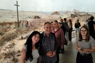 Grupa ludzi w muzeum, w tle panorama krajobrazu z krzyżem i ruinami.