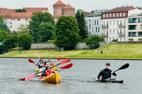 Grupa osób w kajakach na rzece, w tle zamek i zabudowania miejskie.
