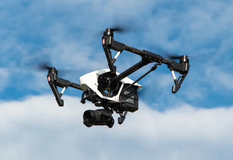 Biało-czarny dron z dużym obiektywem aparatu unosi się w powietrzu na tle błękitnego niezachmurzonego nieba 