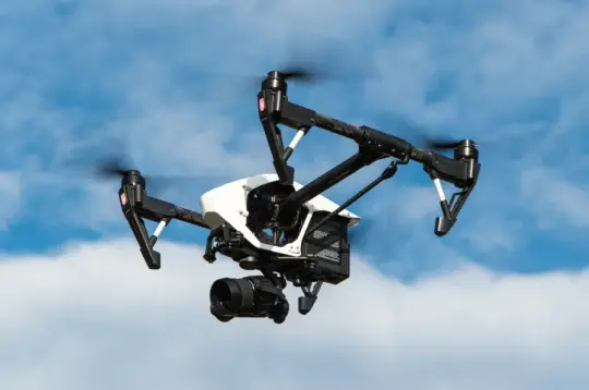 Biało-czarny dron z dużym obiektywem aparatu unosi się w powietrzu na tle błękitnego niezachmurzonego nieba 