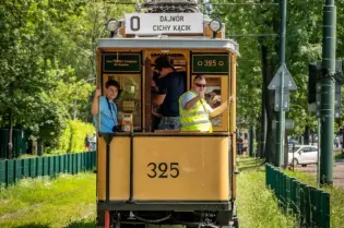 Tramwaj na torach z pasażerami, otoczony zielenią drzew.