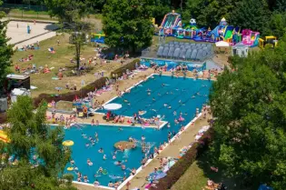 Basen z atrakcjami wodnymi, zjeżdżalniami i plażą, pełen ludzi w letniej scenerii.