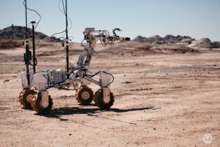 Na różowym pustynnym podłożu stoi robotyczny srebrny łazik planetarny. 