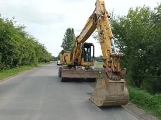 Żółty koparko-ładowarka na drodze, otoczona zielenią.