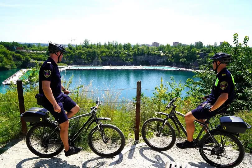 Dwaj policjanci na rowerach obserwują zbiornik wodny otoczony zielenią.