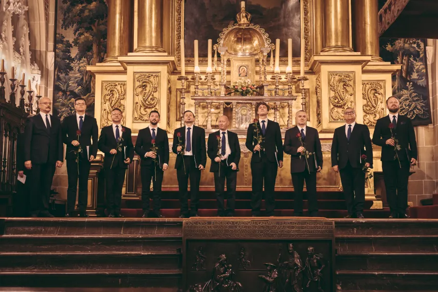 10 mężczyzn ubranych w czarne garnitury stoi na stopniach zabytkowego kościelnego ołtarza. Każdy z nich trzyma w ręku jedną czerwoną różę. 