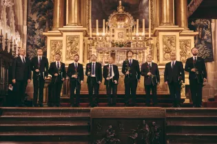 10 mężczyzn ubranych w czarne garnitury stoi na stopniach zabytkowego kościelnego ołtarza. Każdy z nich trzyma w ręku jedną czerwoną różę. 