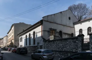 Budynek Synagogi Kupa na krakowskim Kazimierzu w czasach współczesnych. Widoczny budynek o charakterystycznej bryle bożnicy fragment ulicy z zaparkowanymi samochodami  