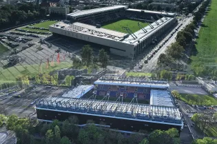 Widok na stadion i otaczający go teren, z zielonymi przestrzeniami i budynkami w tle.