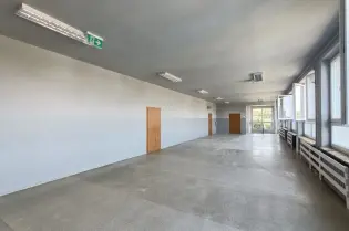 Pusta sala z jasnymi ścianami, oknami i drzwiami, podłoga w kolorze szarym.