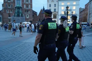 Grupa strażników miejskich patrolujących rynek w mieście, w tle tłum ludzi i zabytkowe budynki.