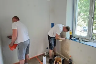 Dwaj mężczyźni malują ściany w pokoju, jeden z nich nakłada farbę, drugi przygotowuje narzędzia.