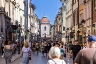 liczne osoby idą pojedynczo ulicą Floriańską w Krakowie. Tłum przechodniów widać od tyłu na tle oddalonej ceglanej Bramy Floriańskiej.    