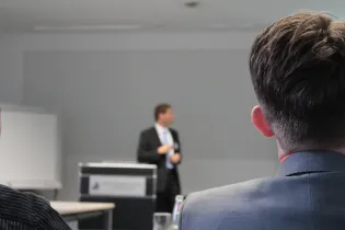 Uczestnicy wykładu słuchają prelegenta stojącego przy mównicy w sali konferencyjnej.