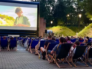 Kino plenerowe z widownią siedzącą na leżakach, wyświetlany film na dużym ekranie.