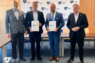 Czterech mężczyzn w garniturach trzyma dokumenty, stojąc w biurze z logo Kraków Airport w tle.