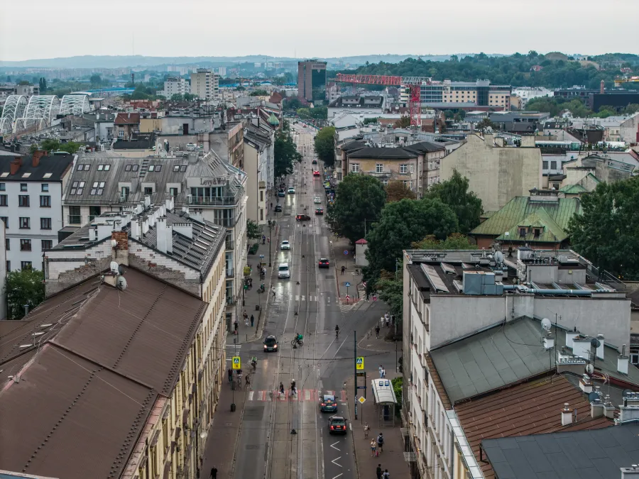 Widok na ulicę z zabudową miejską, samochody i zieleń w tle.