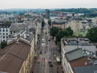 Widok na ulicę z zabudową miejską, samochody i zieleń w tle.