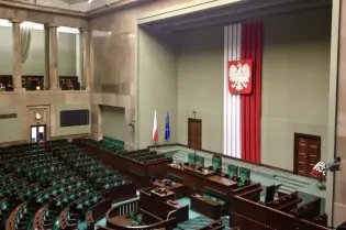 Wnętrze sali obrad Sejmu z flagami i godłem Polski na tle ściany.