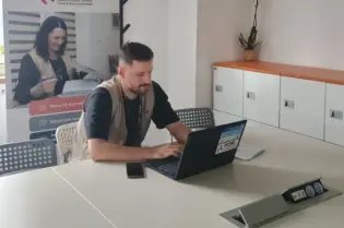 Zdjęcie przedstawia mężczyznę pracującego przy laptopie przy stole. W tle widoczny jest roll-up dotyczący wsparcia cudzoziemców w dostępie do edukacji oraz integracji społecznej i zawodowej w Polsce.