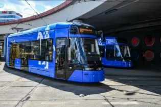 Niebieski tramwaj na skrzyżowaniu, w tle drugi tramwaj oraz nowoczesne budynki.