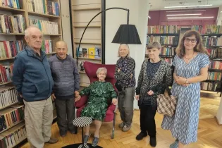 Grupa sześciu osób w bibliotece, w tym starsza kobieta siedząca na fioletowym fotelu.