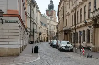 Ulica z kamienicami i wieżą zegarową w tle. Parkujące samochody na jezdni.