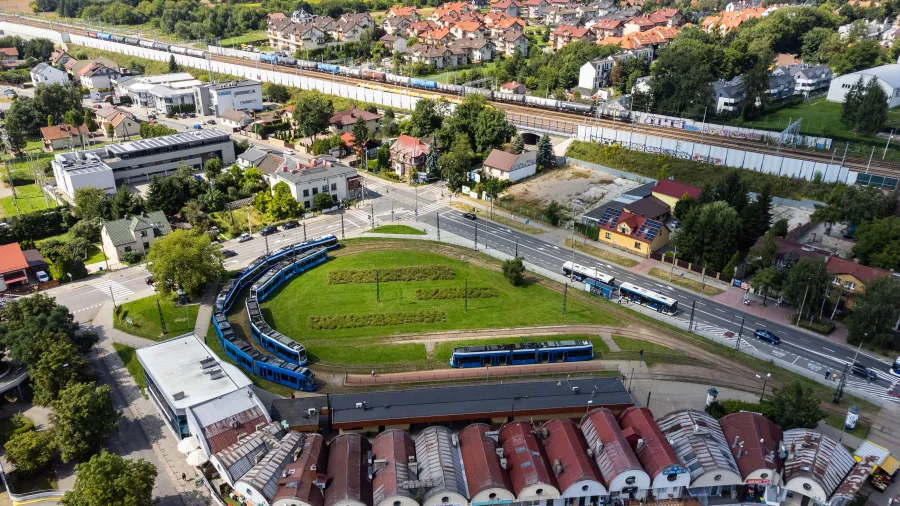 Tramwaj na pętli w otoczeniu zieleni i zabudowy miejskiej.