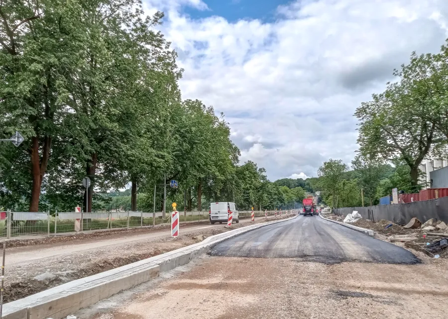 Prace drogowe w otoczeniu drzew, z widoczną nową nawierzchnią i pojazdami budowlanymi.