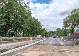 Prace drogowe w otoczeniu drzew, z widoczną nową nawierzchnią i pojazdami budowlanymi.