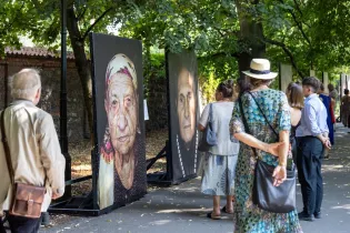 Wystawa portretów ludzi w plenerze, zwiedzający podziwiają prace artystyczne.