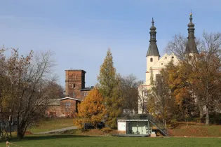 Kościół z wieżami, budynki w tle, jesienne drzewa i zielona trawa.