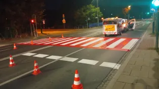 Przejazd dla pieszych malowany na drodze, otoczony pachołkami drogowymi.