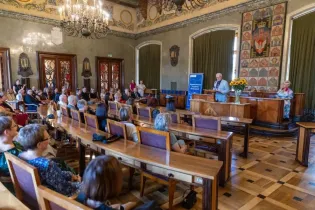 Spotkanie w sali konferencyjnej z mówcą i słuchaczami, dekoracje na ścianach oraz kwiaty na stole.
