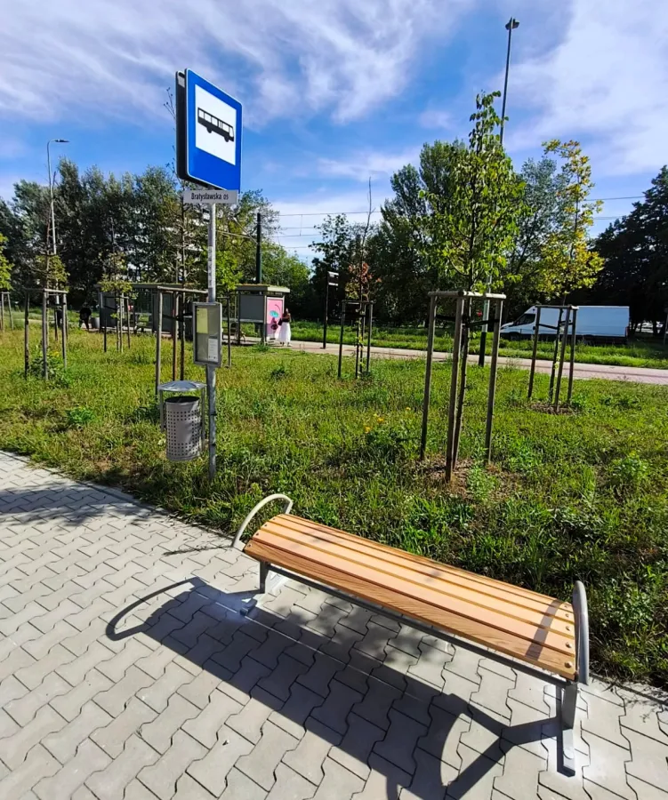 Ławka na przystanku autobusowym z zielenią w tle i niebieskim znakiem przystanku.