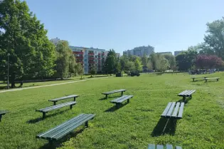 Zielony park z ławkami, drzewami i budynkami w tle. Słoneczna pogoda.
