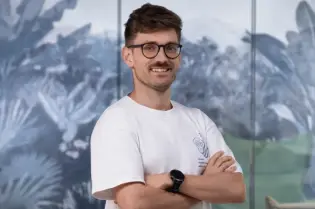 Mężczyzna w okularach, ubrany w biały t-shirt i brązowe spodnie, stoi z założonymi rękami.