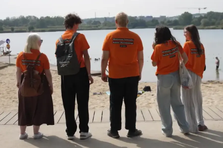 Grupa osób w pomarańczowych koszulkach stoi nad wodą, z widokiem na plażę i kąpiących się.