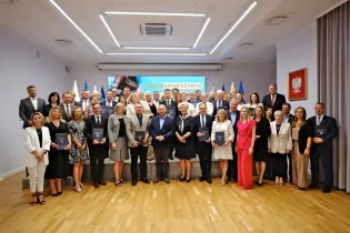Grupa osób pozująca do zdjęcia na tle flag i prezentacji, trzymająca dokumenty.