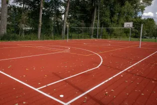 Boisko sportowe z czerwoną nawierzchnią, linie do koszykówki i piłki nożnej, otoczone drzewami.