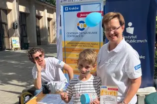 Zdjęcie ze stoiska Centrum Obywatelskiego Centrum C 10 i Fundacji Instytut Polityk Publicznych podczas wydarzenia plenerowego. Przy stoisku stoją dwie osoby dorosłe i chłopiec.