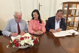 Trzy osoby podpisujące dokumenty przy stole, z kwiatami w wazonie.